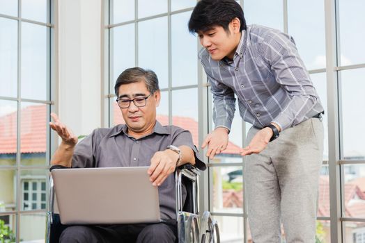 Asian senior disabled businessman in a wheelchair with laptop computer discuss together with team in office. Old father man sitting wheelchair and his son talking video calls conference on laptop