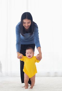 Asian little baby girl taking first steps learning to walk with mom help support the cute unstable walking toddler at home in bedroom. Happy family first steps parenthood concept
