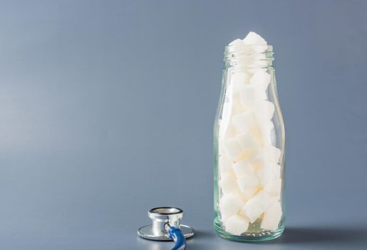 Glass bottle full of white sugar cube sweet food ingredient and doctor stethoscope isolated gray background, health high blood risk of diabetes and calorie intake concept and unhealthy drink