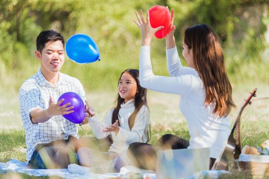 Happy Asian young family father, mother and child little girl having fun and enjoying outdoor sitting on picnic blanket playing balloons at summer garden spring park, Family relaxation concept