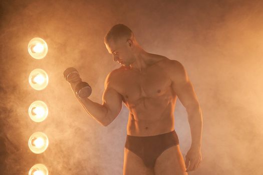 Young athlete pumping dumbbells, practicing biceps training, smoky illuminated background Healthy lifestyle concept. Sport and fitness Lamp ray light