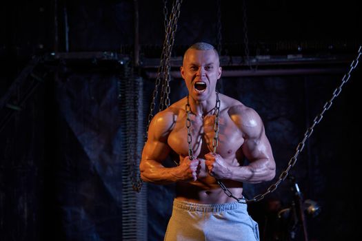 Primal instinct. Angry muscular man screaming at camera and breaking chains on chest, dark background