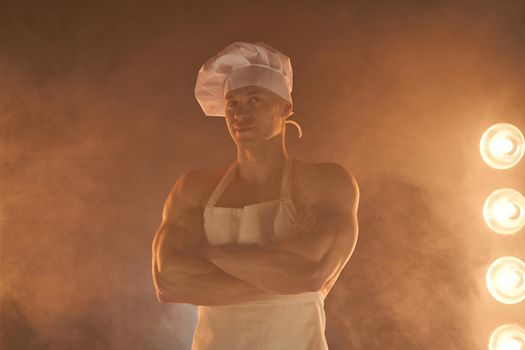 Portrait of muscular chef wearing white apron and chef hat, posing with folded arms on smoky background male housewife. husband in kitchen. brutal butcher.