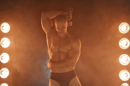 Young athlete pumping dumbbells, practicing triceps training, smoky illuminated background Healthy lifestyle concept. Sport and fitness Lamp ray light