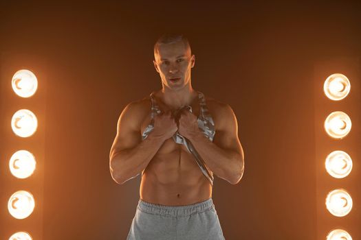 Active lifestyle concept. Professional bodybuilder picks up a t-shirt showing abs muscle and looking at camera, lamps illumination and smoke on background Sport and fitness concept