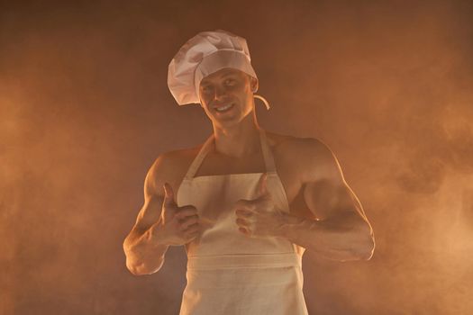 Portrait of muscular chef wearing white apron and chef hat, showing thumb up and smile on smoky background male housewife. husband in kitchen. brutal butcher.