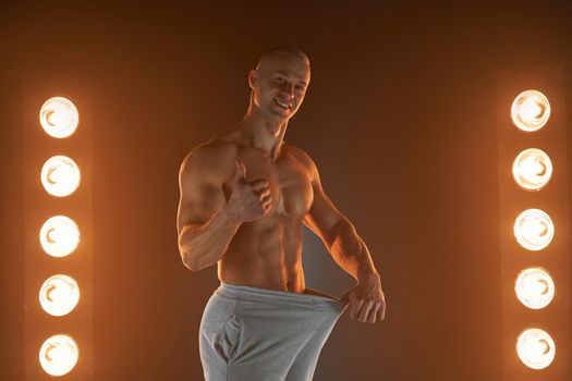 Great male size. Young muscular guy pulling back his pants and showing thumb up gesture, being proud and satisfied with his genitals, lamps illumination on background Weight loss concept Diet