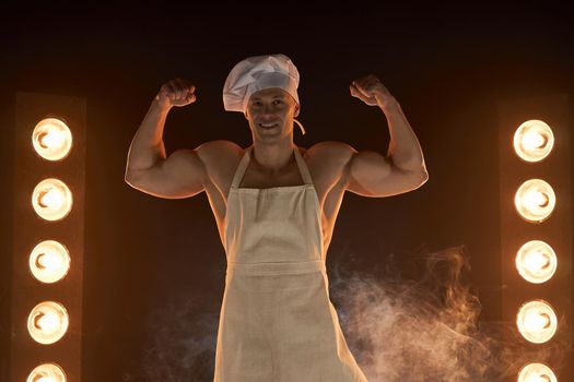 Portrait of muscular chef wearing white apron and chef hat, showing strong biceps muscle on smoky background male housewife. husband in kitchen. brutal butcher.