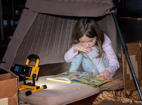First grade girl reading a comic book with a flashlight inside home made tent with boxes and cushions