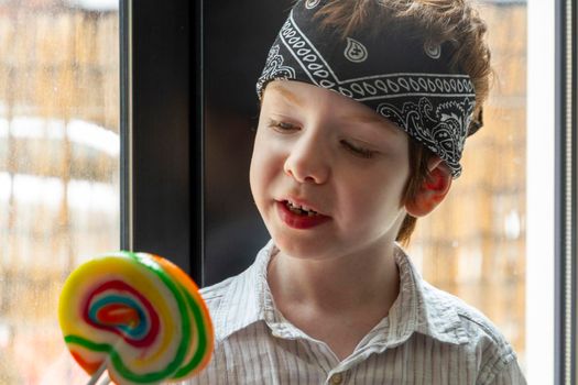 Portrait of a cute elementary age boy, wearing a bandana, looking at a colourful lollipop by a window