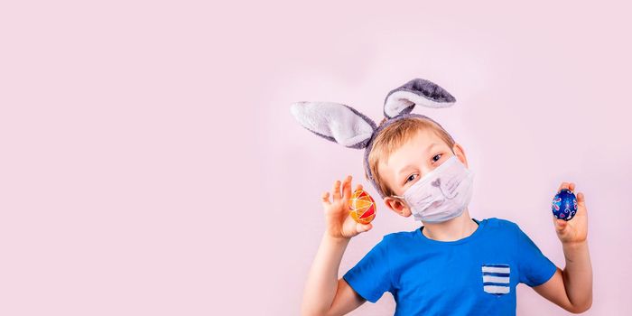 Cute little boy in rabbit bunny ears on head and protective mask with colored eggs on pink background. Cheerful smiling happy child. Covid Easter holiday banner