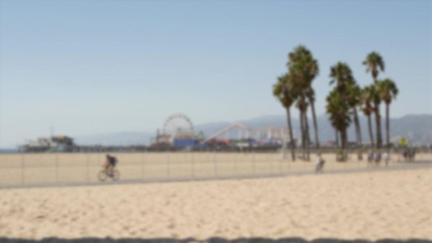 California beach aesthetic, people ride cycles on a bicycle path. Blurred, defocused background. Amusement park on pier and palms in Santa Monica american pacific ocean resort, Los Angeles CA USA.