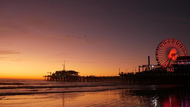 Twilight waves against classic illuminated ferris wheel, amusement park on pier in Santa Monica pacific ocean beach resort. Summertime iconic symbol of California glowing in dusk, Los Angeles, CA USA