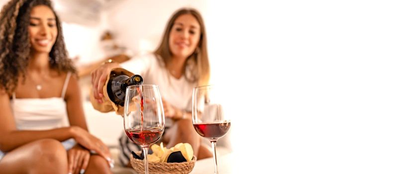 Two happy female friends celebrating at home pouring red wine in the glasses in selective focus effect and copy space. Young Caucasian woman toasting with her best Hispanic friend drinking alcohol