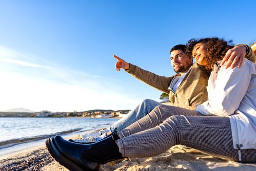 Affectionate young couple in love sitting on seashore near the ocean water next to each others looking at the sunset. Handsome man embracing girlfriend pointing to horizon in winter vacation travel