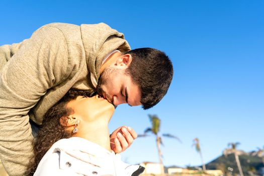 Handsome guy kissing her african-american black girlfriend approaching her mouth from above. Mixed race young couple in love romance vacation scene in sea or ocean resort with copy space in blue sky