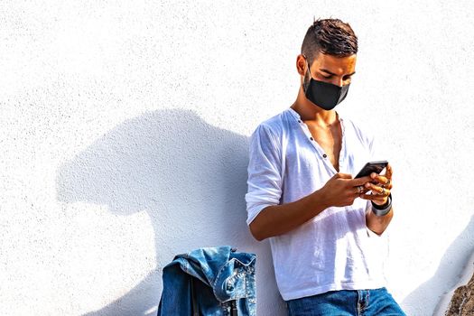 Young solo traveller leaning at a white wall with trolley bag using smartphone chatting online waiting bus or train wearing Coronavirus black protective mask. Modern guy in bright color vivid effect