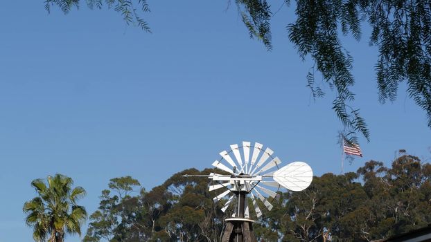 Classic retro windmill, bladed rotor and USA flag against blue sky. Vintage water pump wind turbine, power generator on livestock ranch or agricultural farm. Rural symbol of wild west, rustic suburb.