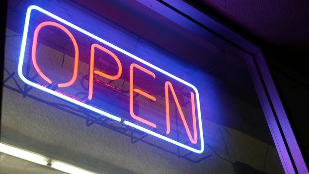 Open neon sign glowing in the dark. Vivid retro styled text at entrance on glass window. Colorful electric banner selective focus close up. Light bulbs radiance at night. Shiny illuminated lettering.