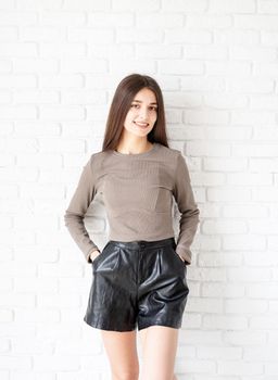 Three quarters length portrait of beautiful smiling brunette woman with long hair wearing brown shirt and black leather shorts, on white brick wall background, hands in pockets