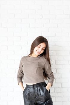 Three quarters length portrait of beautiful smiling brunette woman with long hair wearing brown shirt and black leather shorts, on white brick wall background, hands in pockets