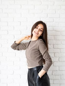 Three quarters length portrait of beautiful smiling brunette woman with long hair wearing brown shirt and black leather shorts, on white brick wall background