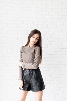 Three quarters length portrait of beautiful smiling brunette woman with long hair wearing brown shirt and black leather shorts, on white brick wall background