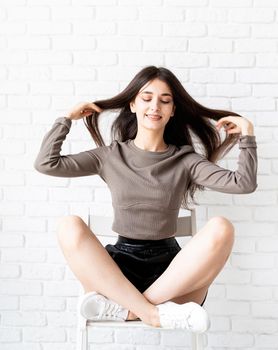 portrait of beautiful smiling brunette woman with long hair wearing brown shirt and black leather shorts sitting on white brick wall background