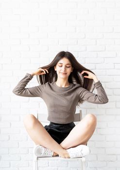 portrait of beautiful smiling brunette woman with long hair wearing brown shirt and black leather shorts sitting on white brick wall background