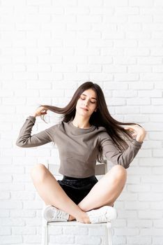 portrait of beautiful smiling brunette woman with long hair wearing brown shirt and black leather shorts sitting on white brick wall background