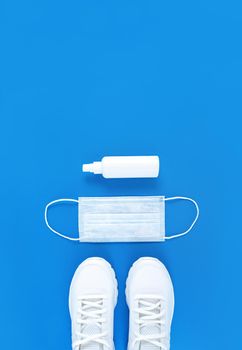 White medical mask, trainers, and hand sanitizer on a blue background. Monochrome vertical flat lay.