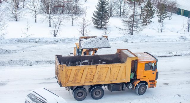 Big yellow tractor cleans up snow from the road and loads it into the truck. Cleaning and cleaning of roads in the city from snow in winter