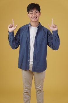 portrait of happy young asian man dressed casually pointing finger up at copyspace. yellow studio background