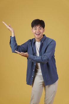 portrait of happy handsome young asian man dressed casually presenting with open palms gesture at copyspace. yellow studio background