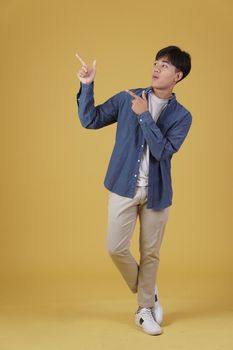 portrait of happy handsome young asian man dressed casually pointing fingers at copyspace isolated on yellow studio background