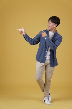 portrait of happy handsome young asian man dressed casually pointing fingers at copyspace isolated on yellow studio background