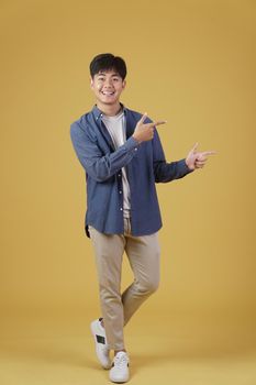 portrait of happy handsome young asian man dressed casually pointing fingers at copyspace isolated on yellow studio background