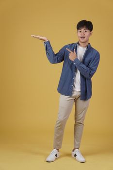 portrait of happy smiling young asian man dressed casually pointing finger to empty open hand isolated on yellow background