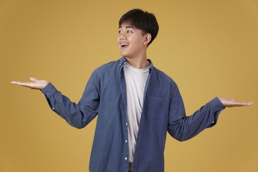 portrait of excited happy handsome young asian man dressed casually smiling with open hand gesture isolated on yellow studio background