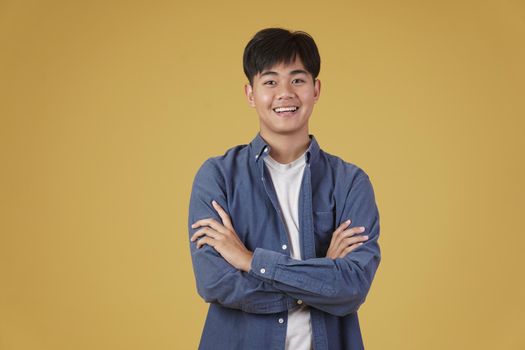 portrait of happy handsome young asian man dressed casually with folded crossed arms gesture isolated on yellow studio background