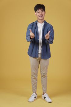 portrait of smiling positive young asian man dressed casually with thumb up gesture approving expression isolated on yellow studio background