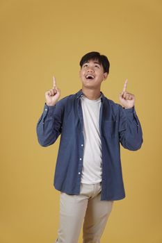 portrait of happy young asian man dressed casually pointing finger up at copyspace. yellow studio background