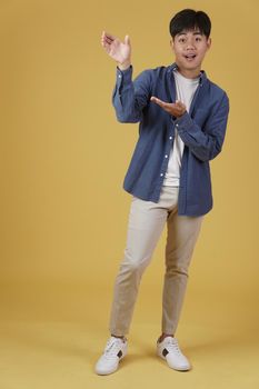 portrait of happy handsome young asian man dressed casually presenting with open palm hand gesture at copyspace isolated on yellow studio background