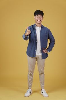 portrait of smiling positive young asian man dressed casually with thumb up gesture approving expression isolated on yellow studio background