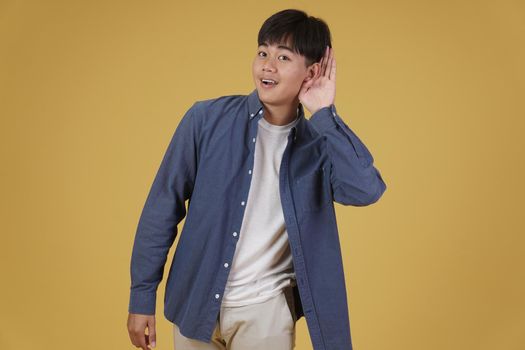 portrait of cheerful happy rejoicing young asian man dressed casually putting hand on ear listening to something isolated on yellow studio background