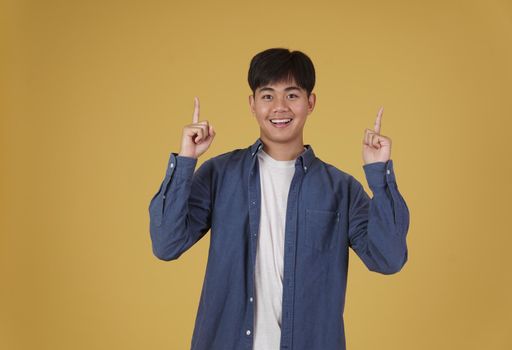 portrait of happy young asian man dressed casually pointing finger up at copyspace. yellow studio background