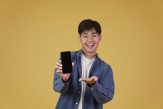 portrait of happy young asian man dressed casually smiling showing smartphone blank white screen isolated on yellow studio background