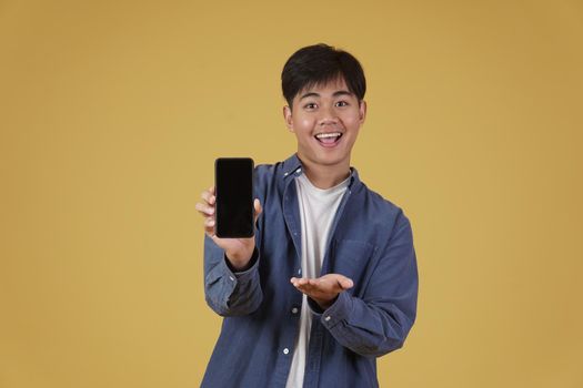 portrait of happy young asian man dressed casually smiling showing smartphone blank white screen isolated on yellow studio background