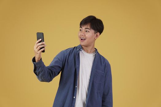portrait of happy surprised excited young asian man dressed casually looking at smartphone isolated on yellow studio background