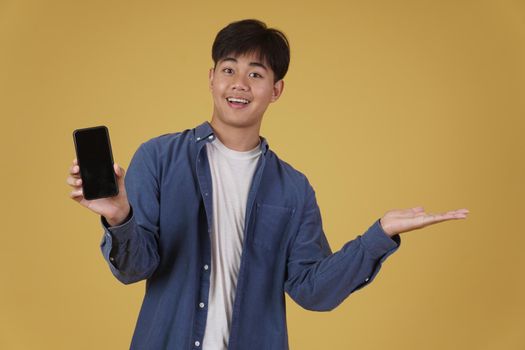 portrait of happy young asian man dressed casually smiling showing smartphone blank white screen isolated on yellow studio background